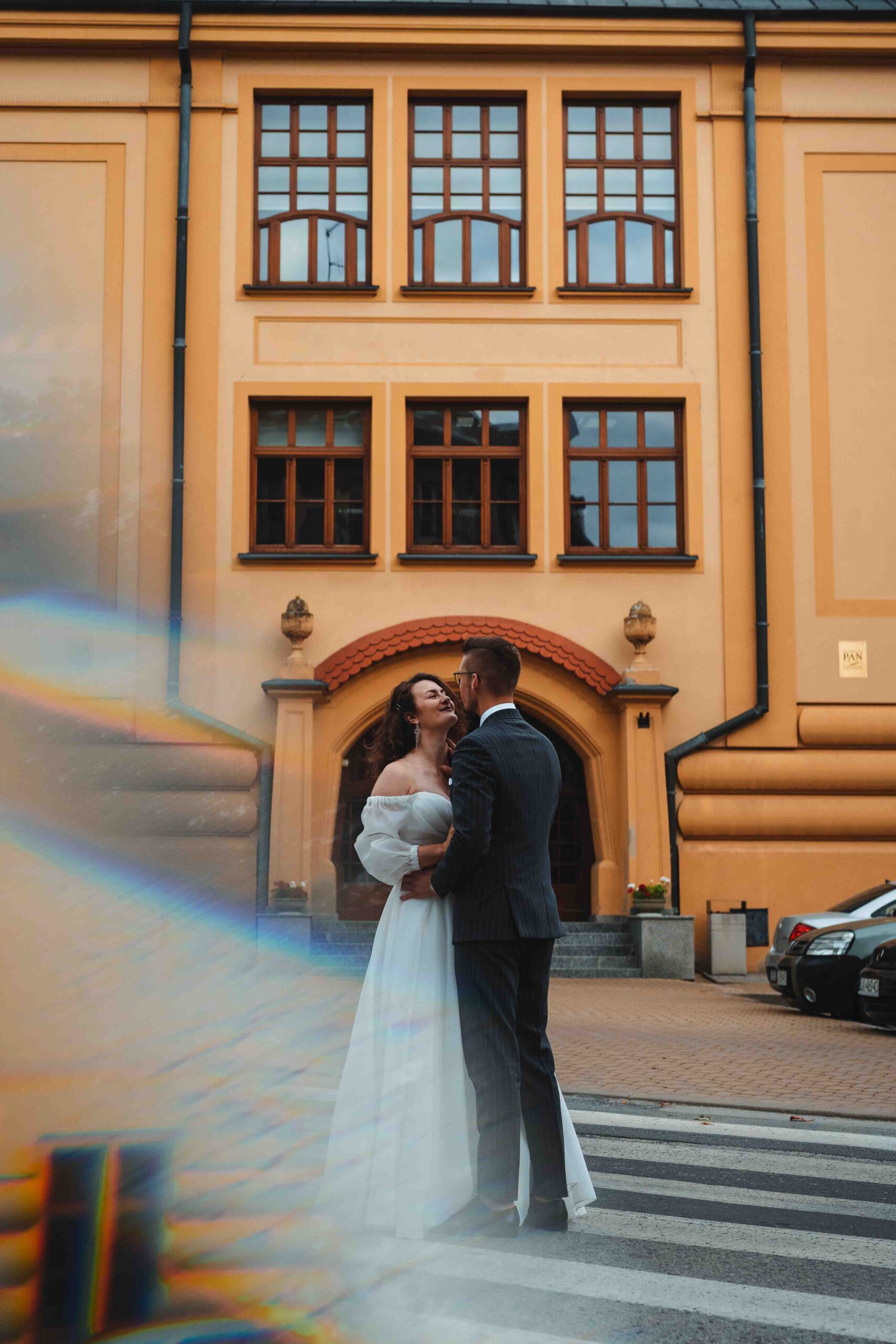 Para Młoda podczas sesji plenerowej przytula się na przejściu dla pieszych przed wysoką kamienicą.