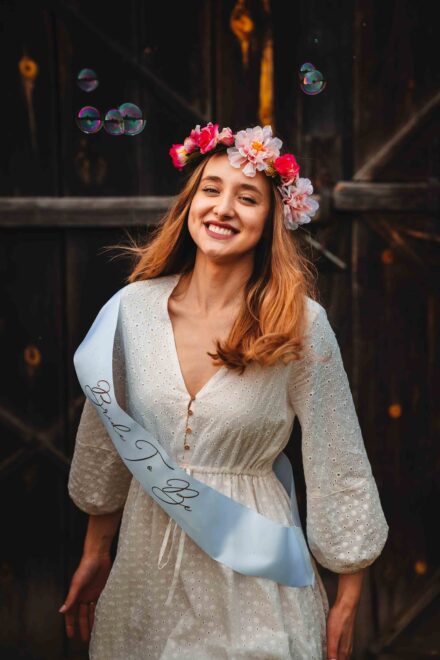 Panna młoda w wianku z kwiatów uśmiecha się na tle drewnianej ściany.
