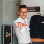 Pan Młody podczas przygotowań. Wskazuje palcem wskazującym w stronę fotografa ślubnego, który robi zdjęcie.