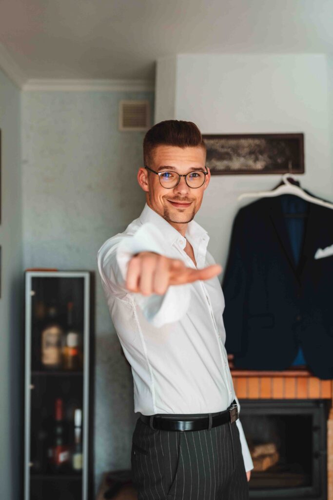 Pan Młody podczas przygotowań. Wskazuje palcem wskazującym w stronę fotografa ślubnego, który robi zdjęcie.