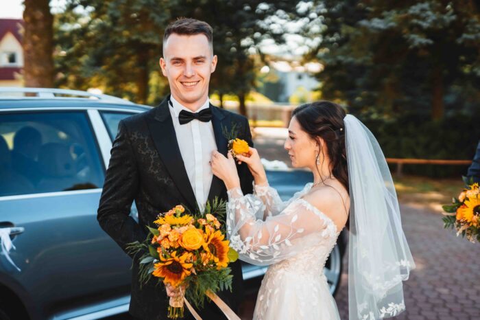 Panna Młoda poprawia butonierkę w marynarce Pana Młodego.