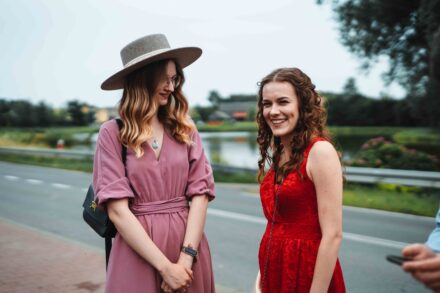 Dwie kobiety czekają przy ulicy na rozpoczęcie ślubnej mszy. W tle widać małe jezioro.