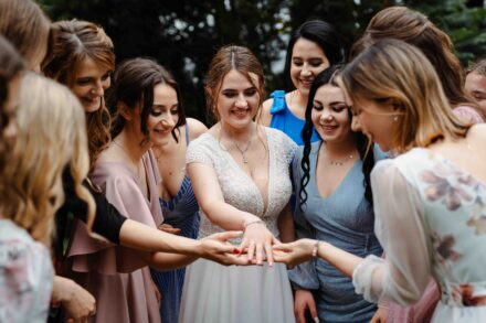 Grupa młodych kobiet ubranych w sukienki podziwia obrączkę Panny Młodej w sukni ślubnej w parku.