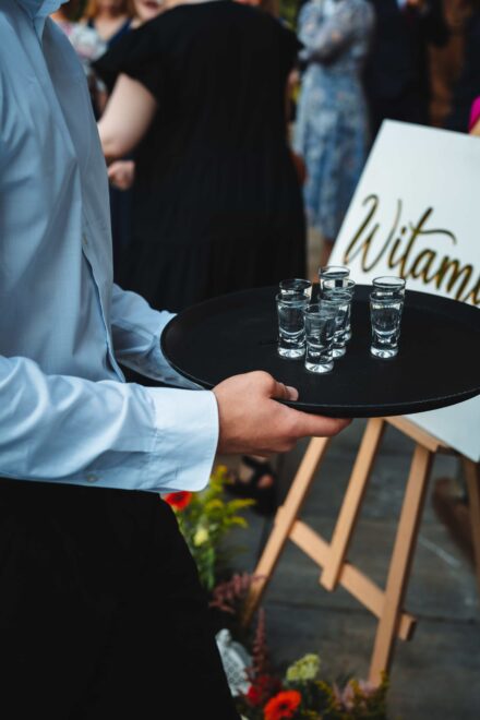 Kelner niesie tacę z ośmioma napełnionymi kieliszkami. W tle widać tablicę powitalną gości.