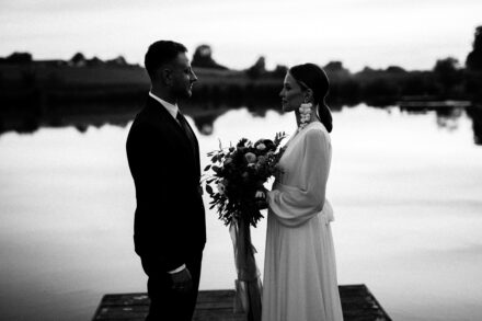 Państwo Młodzi stoją na molo nad jeziorem i spoglądają sobie w oczy. Panna Młoda trzyma bukiet ślubny.