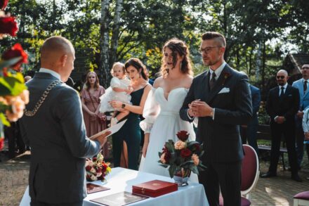 Para Młoda bierze udział w ceremonii ślubu humanistycznego, który odbywa się w dworku. W tle widać gości weselnych i urzędznika.