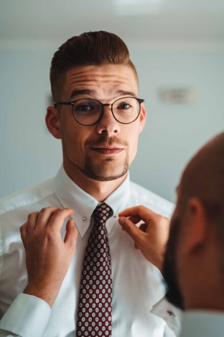 Pan Młody patrzy w kierunku fotografa ślubnego. Trwają przygotowania do ślubu i starszy poprawia spinkę do krawatu Pana Młodego.