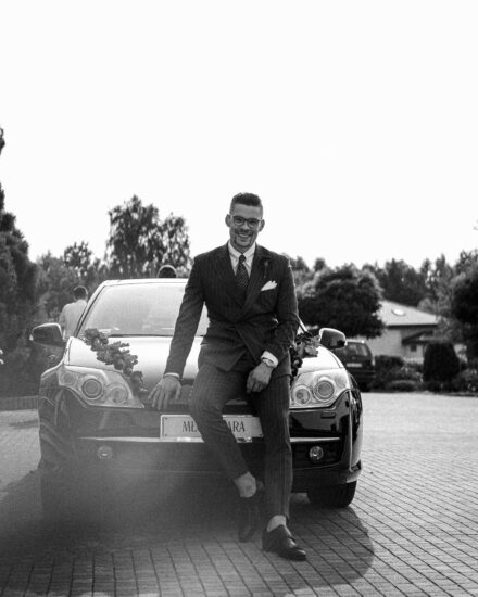 Pan Młody uśmiecha się do fotografa ślubnego i siedzi na masce auta ślubnego.